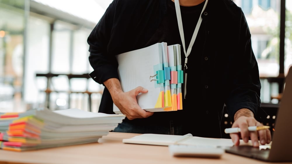 Person med stor bunke dokumentmapper merket med fargerike klistrelapper, jobber på en laptop ved skrivebord fullt av papirarbeid i et moderne kontormiljø.