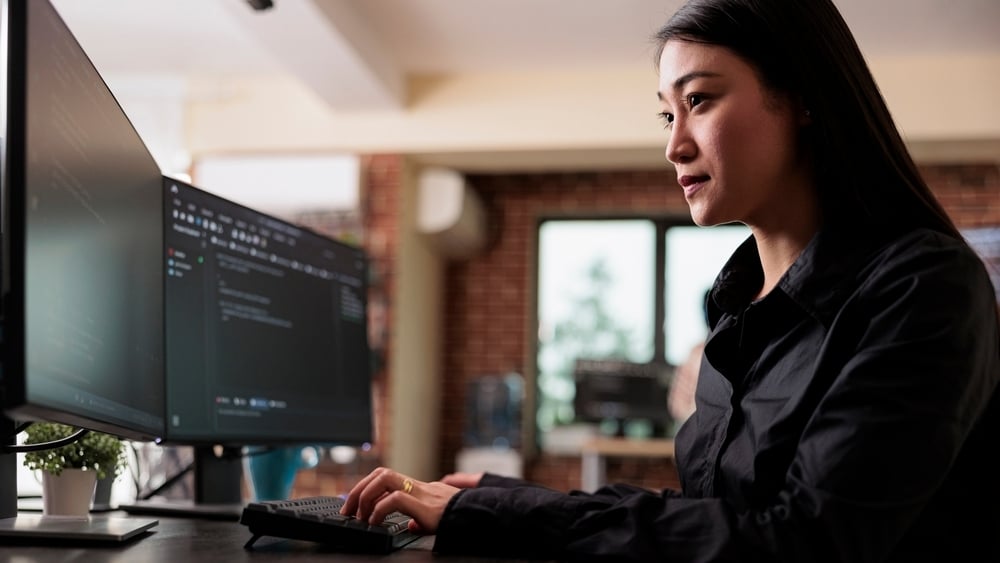 Kvinne sitter ved et skrivebord og jobber på to dataskjermer i et moderne kontormiljø.