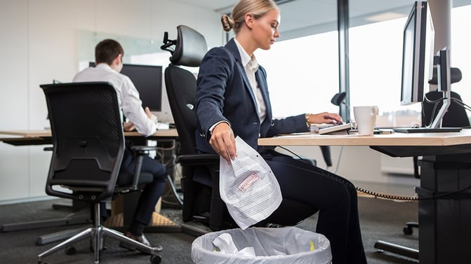 Kvinnelig kontormedarbeider kaster dokumenter i papirkurv. Åpent kontorlandskap med store vindusflater.