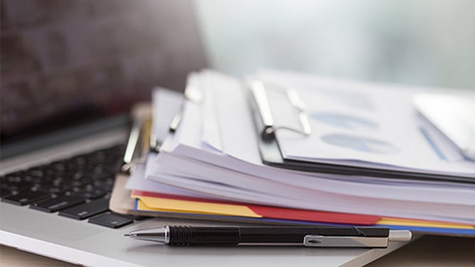 Bunke med dokumenter og permer liggende oppå en åpen laptop, med en penn i forgrunnen – illustrerer behovet for sikker håndtering av papirbasert informasjon på kontor.