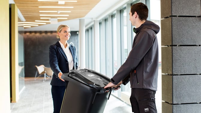 NG Secure-transportør henter avlåst beholder for makulering, hos kontormedarbeider i kontorbygg preget av god belysning fra store vindusflater.